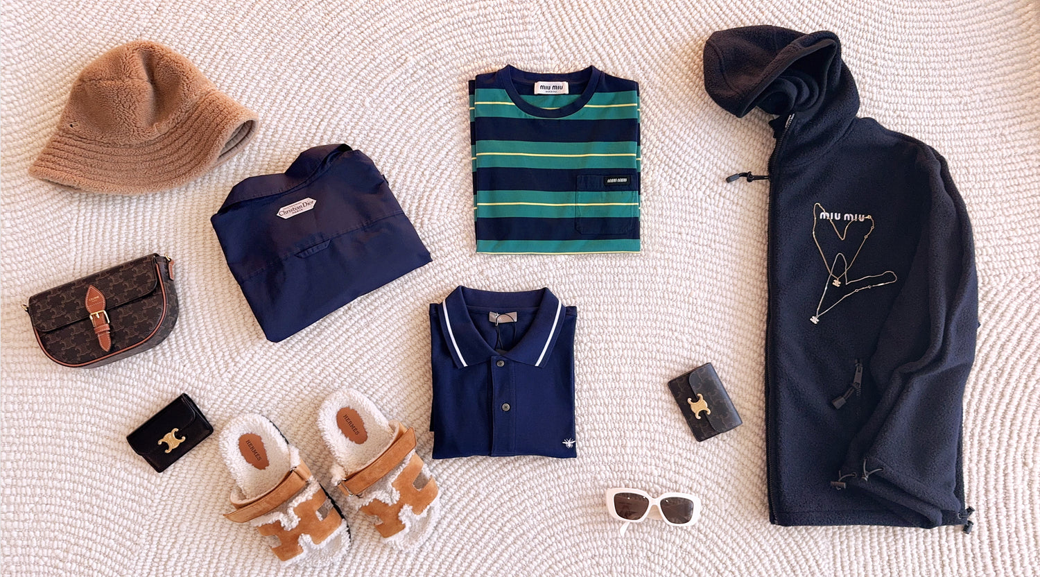 A strategic collection of luxury assets laid out for audit: featuring a Loro Piana teddy bucket hat in the top left, balanced by Hermès teddy shearling sandals. A Dior bee-embroidered polo shirt sits below a reverse-folded Dior jacket, showcasing its iconic interior label. A Miu Miu nautical striped knit and a heavy navy fleece hoodie are paired with a Celine Triomphe bag, card holders, and a classic Celine necklace elegantly draped over the Miu Miu branding. Natural light highlights the authentic Sourcing